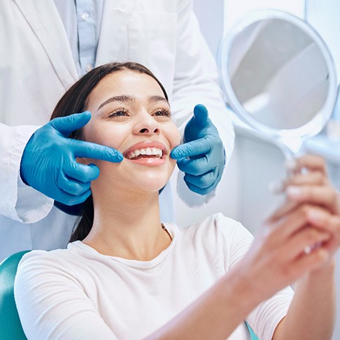 Blue-gloved hands touching corners of woman’s smile as she looks at reflection in mirror