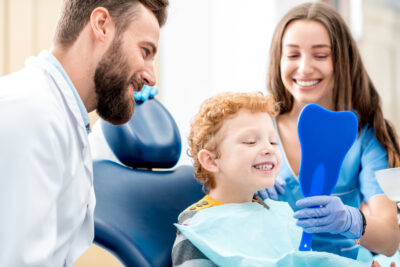 Young boy looking at the mirror with toothy smile sitting on the chair with dentist and assistant at the dental office.