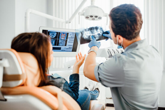 A dentist shows a dental X-ray to a patient in a clinic, discussing the image on a monitor while the patient sits in a dental chair.