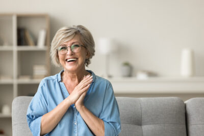 A woman sitting on a couch, smiling and holding her hands together in a joyful manner.