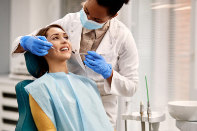 Dentist wearing gloves and a mask examines a smiling patient’s teeth in a dental clinic. The patient is seated in a dental chair and wearing a bib.