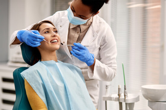Dentist wearing gloves and a mask examines a smiling patient’s teeth in a dental clinic. The patient is seated in a dental chair and wearing a bib.